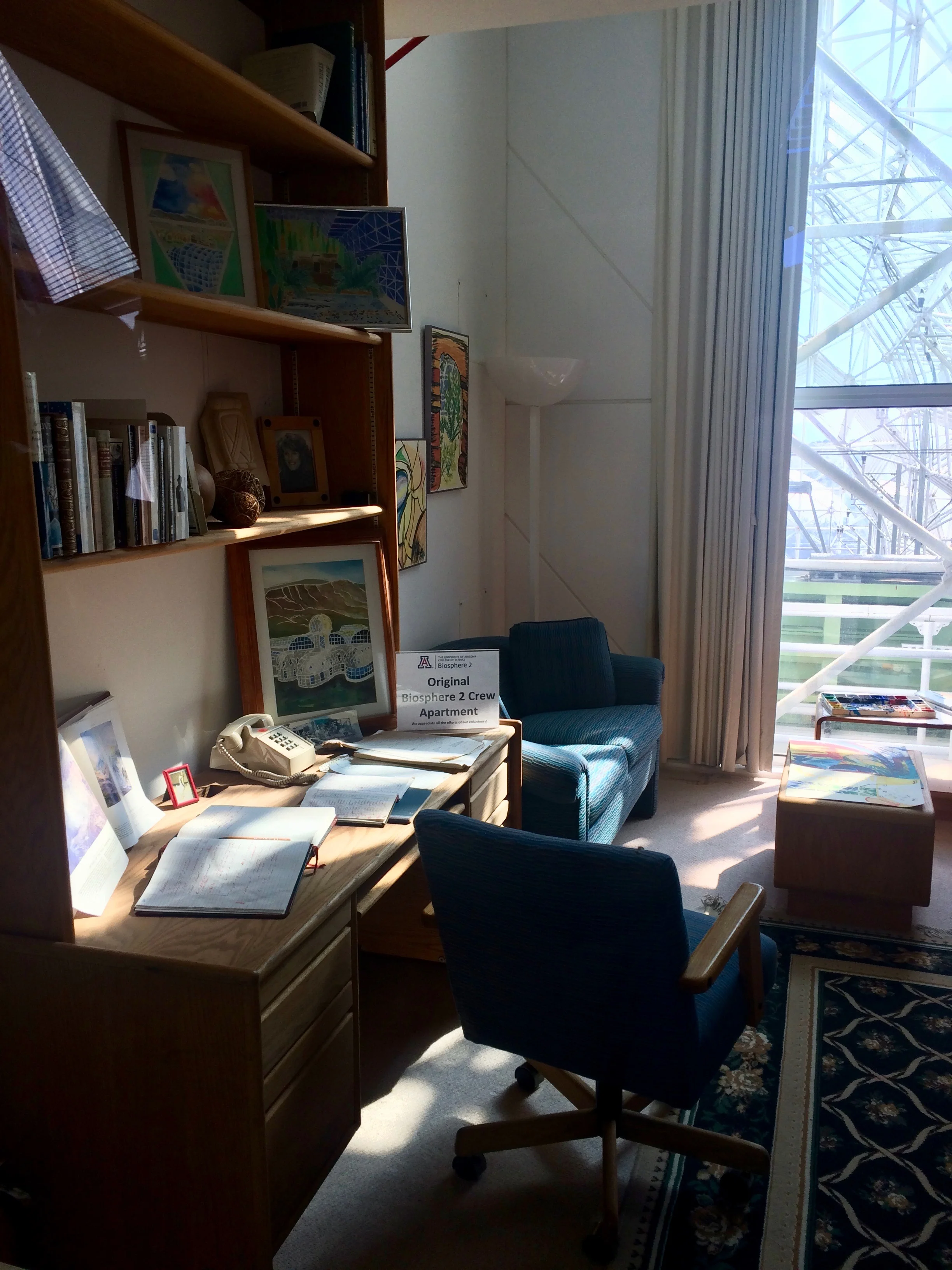 A furnished Biosphere 2 crew apartment with a desk, armchairs, books, and artwork.