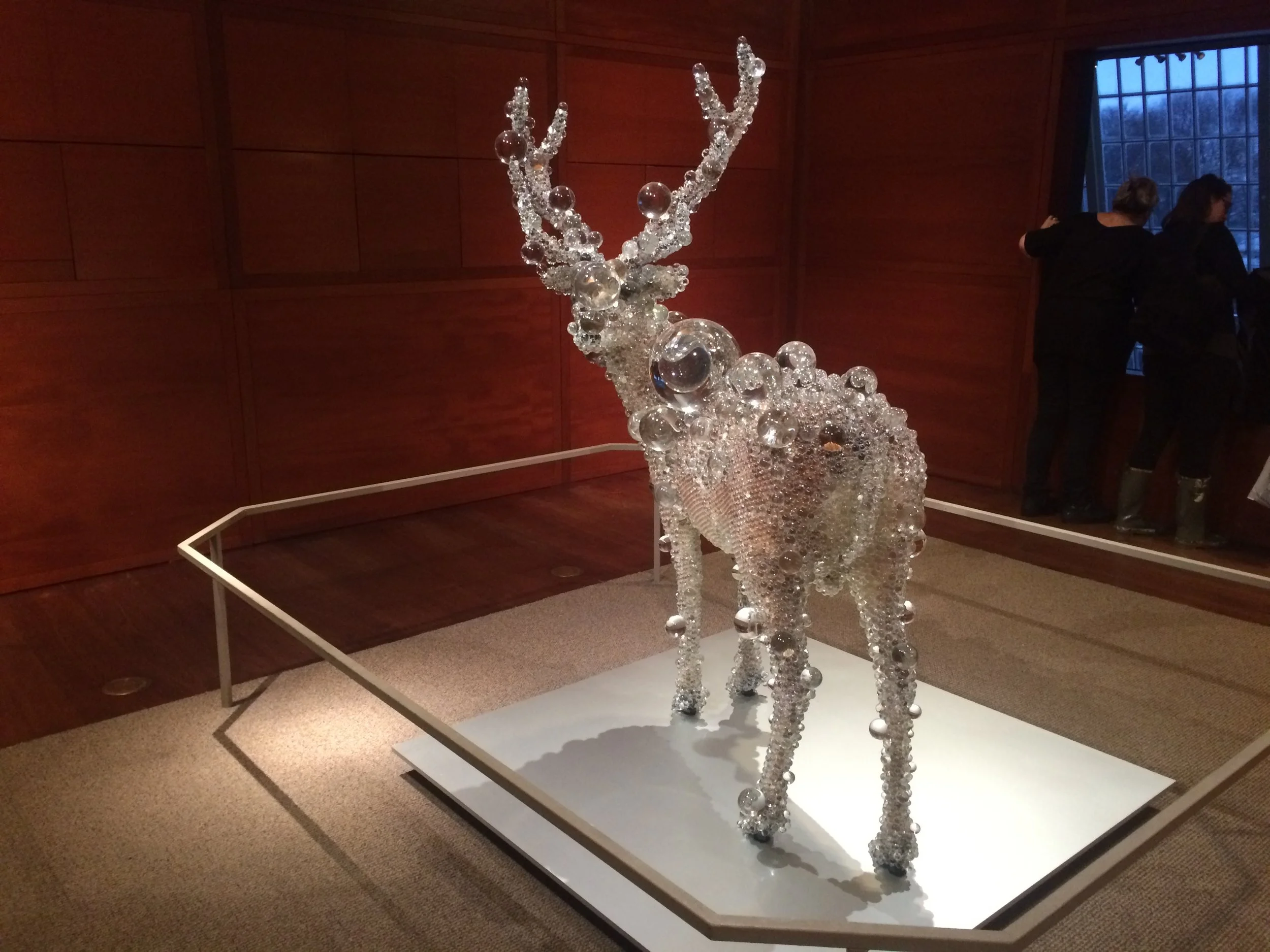 Deer sculpture covered in hundreds of clear glass bubbles, installed against a red museum wall.