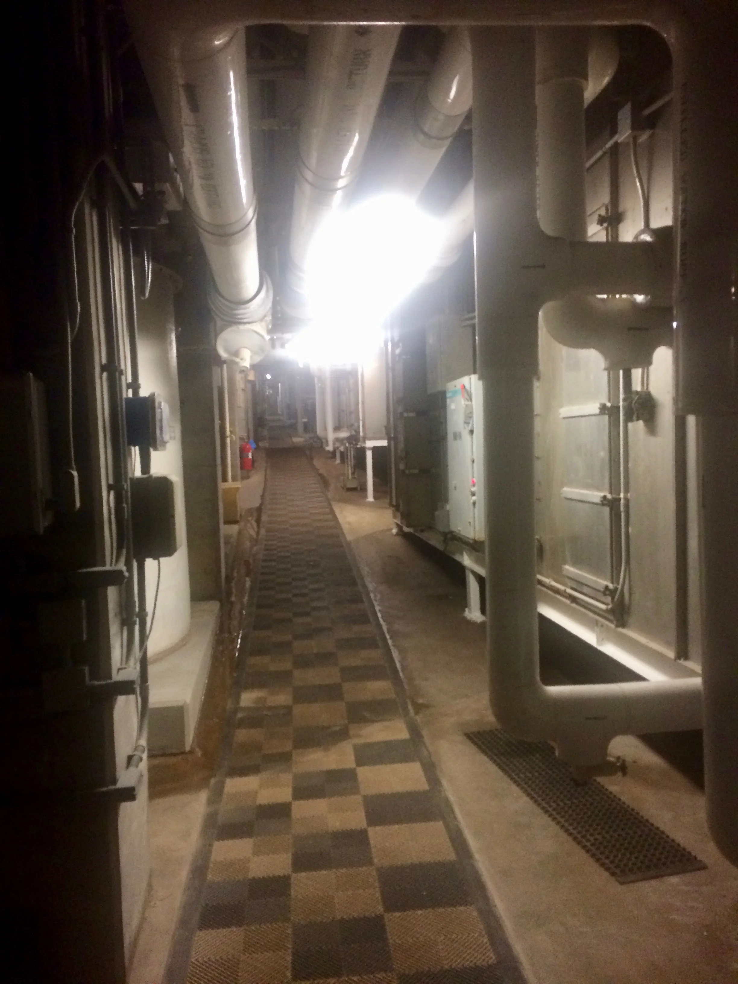 A dim technosphere corridor lined with pipes, ducts, and mechanical equipment.